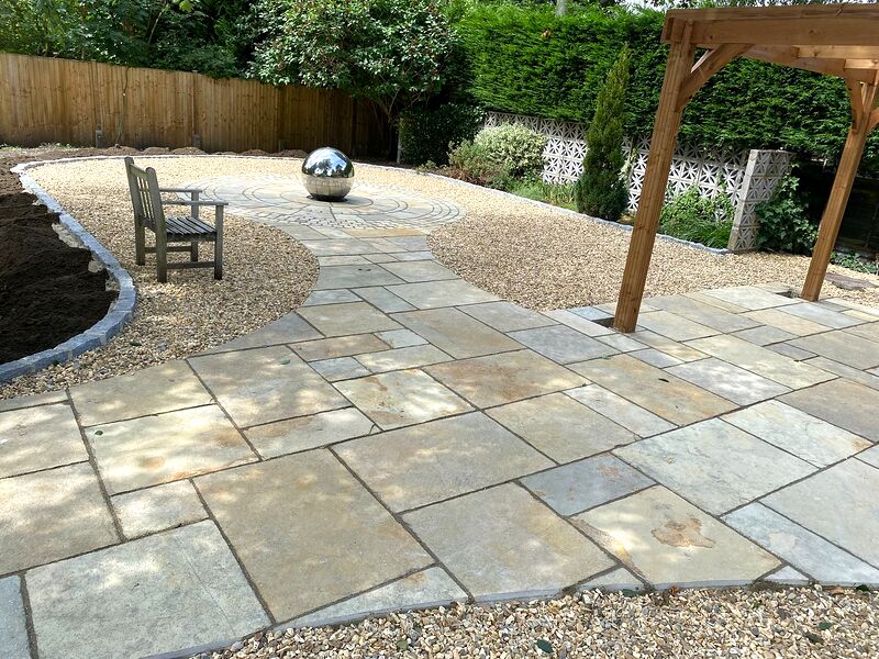 Wide view of a sandstone paved garden with granite-edged gravel, a bench and a sphere water feature