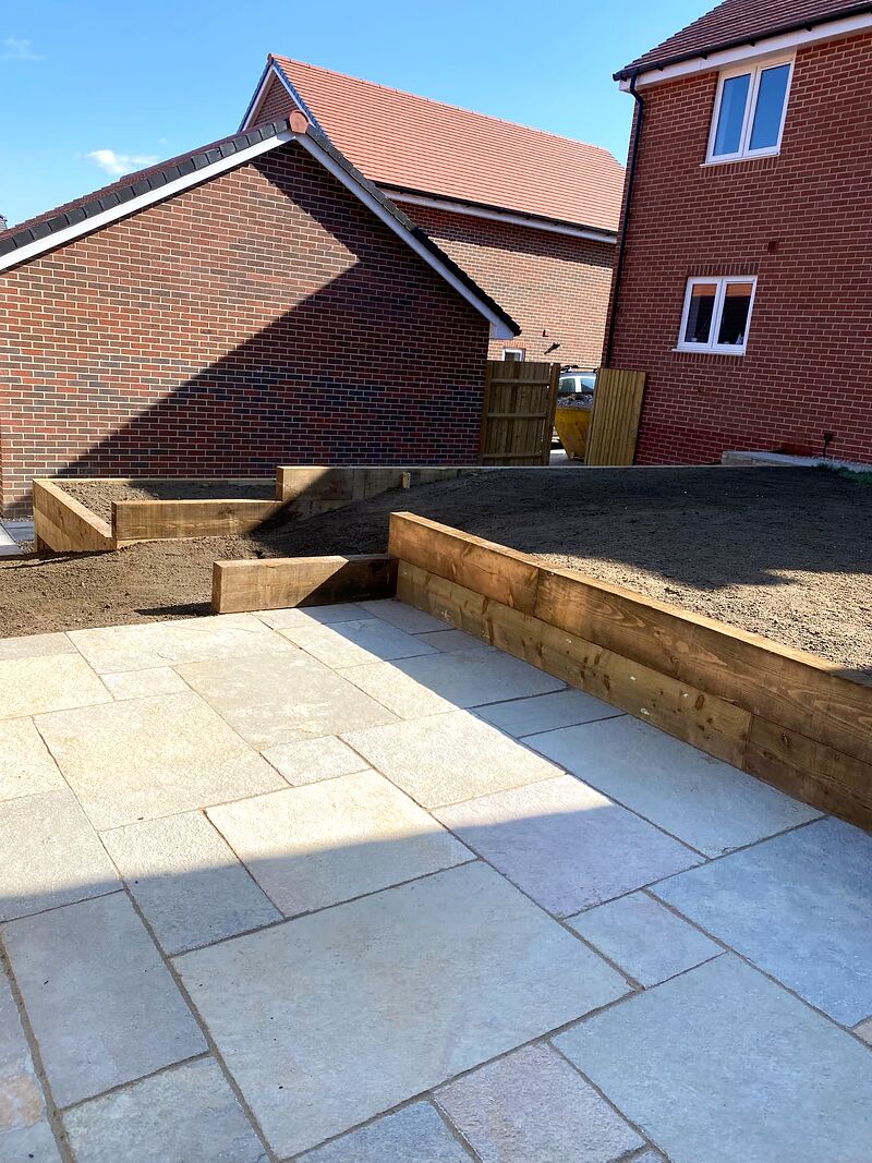 Pale sawn sandstone patio with timber sleeper raised beds beside a new brick house
