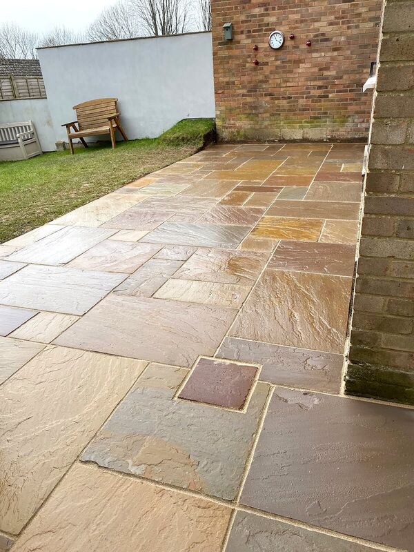 Sandstone patio laid beside a red brick house wall with a lawn and bench beyond